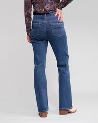 Someone is standing with one hand on their hip, facing away from the camera in Vassalli Theory Jean - Dark Blue Brushed, a patterned long-sleeve shirt, and beige heeled shoes against a plain background.