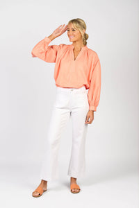 A woman stands against a white background, smiling and raising her right hand in a casual salute. She wears Vassalli’s Daily Jean - White Denim, a peach long-sleeve blouse, tan sandals, and has her hair tied back in a low ponytail.