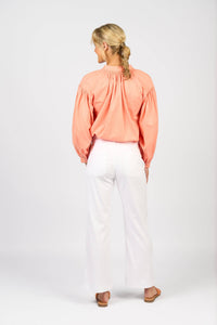A woman with blonde braided hair faces away, wearing a loose coral blouse and Vassalli's Daily Jean - White Denim, featuring visible back pockets and a straight leg. She pairs them with flat sandals against a plain white backdrop.