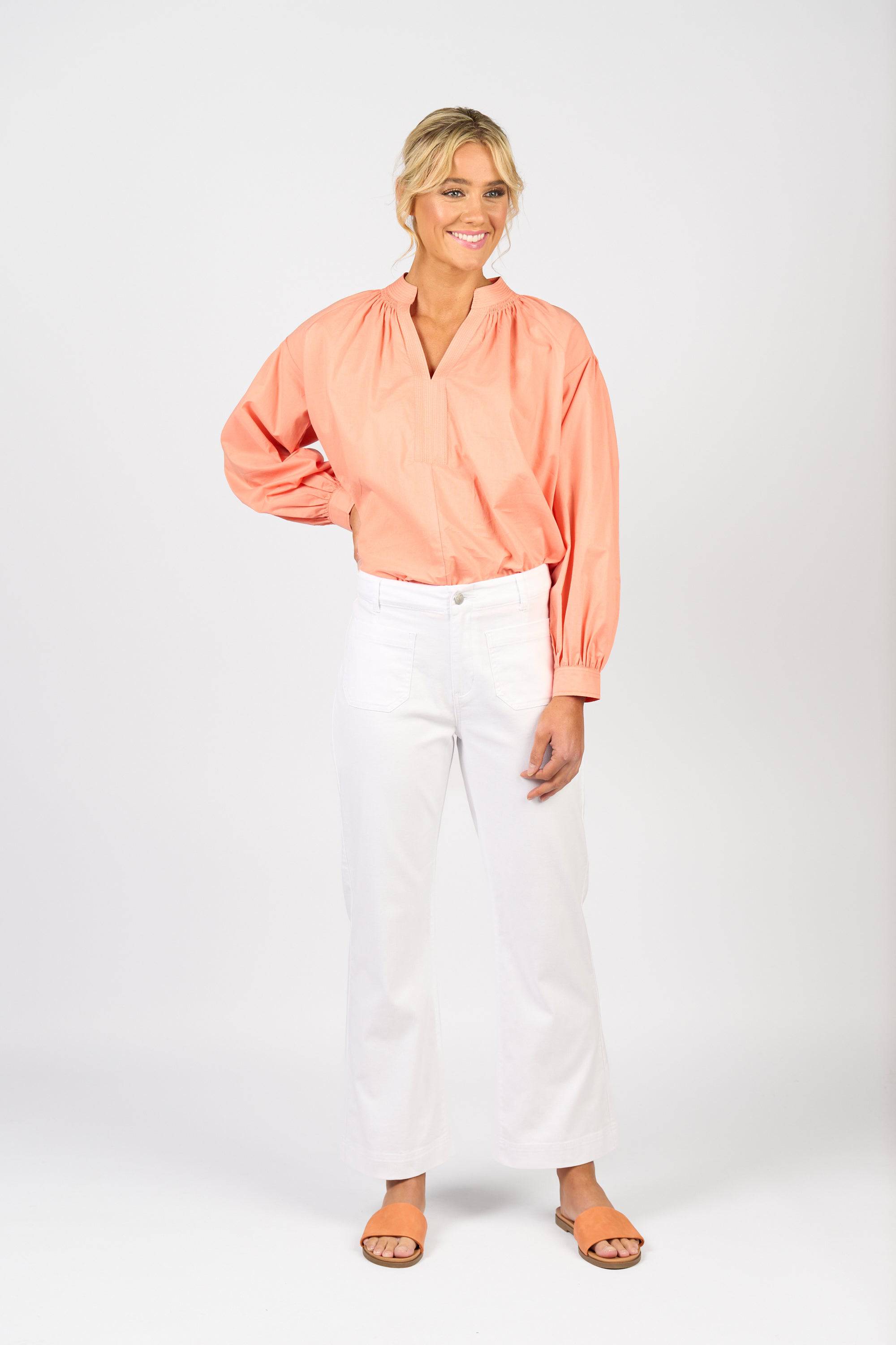 A woman with blonde hair tied back smiles against a white background, wearing Vassalli's Daily Jean in White Denim (ankle-grazer straight leg), a coral long-sleeve blouse, and tan sandals, posing with one hand on her hip.