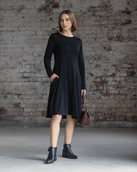 A woman with short brown hair and a neutral expression stands before a textured brick wall, wearing the Vassalli Lotus Merino Dress in black with black ankle boots and holding a burgundy handbag.