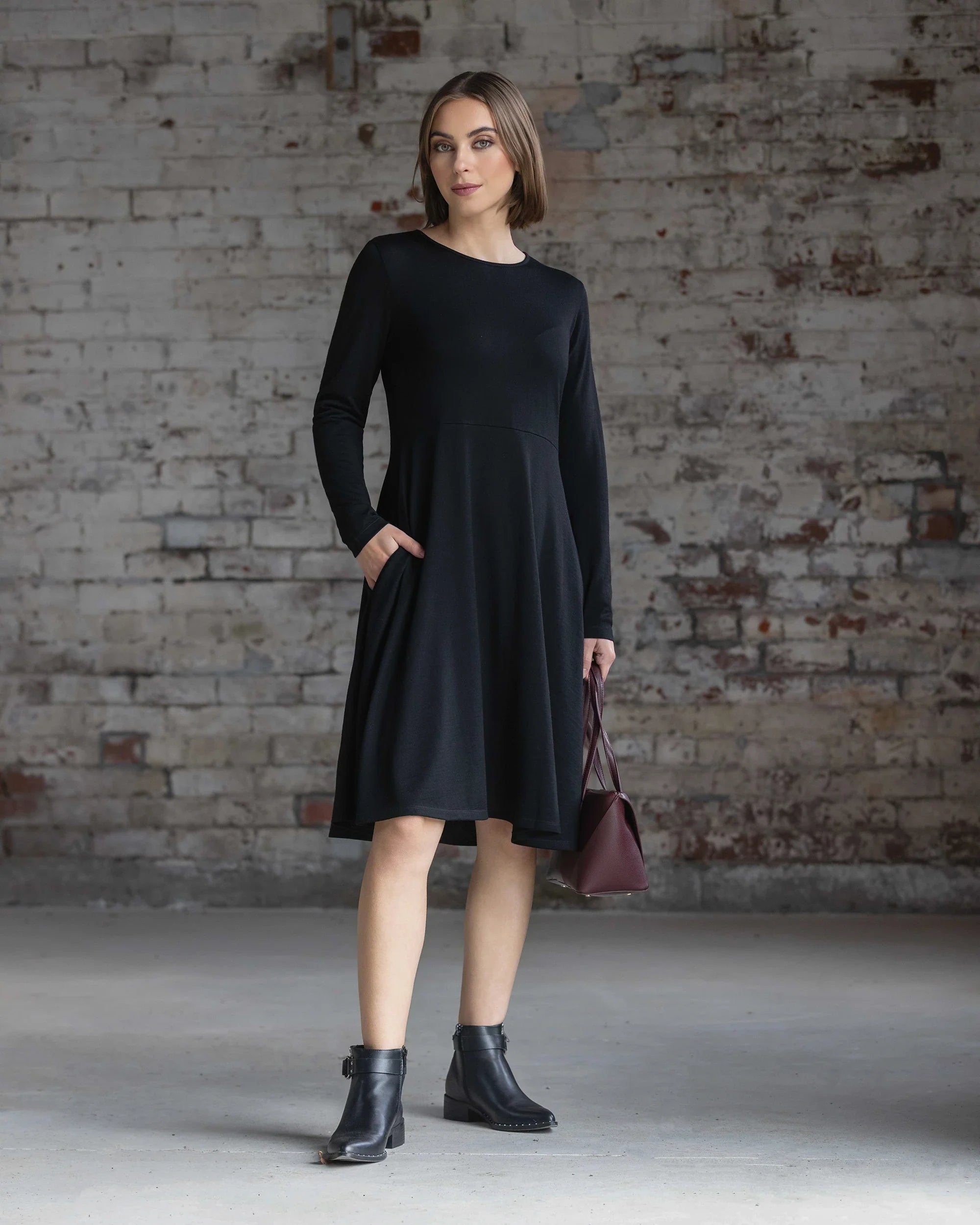 A woman with short brown hair and a neutral expression stands before a textured brick wall, wearing the Vassalli Lotus Merino Dress in black with black ankle boots and holding a burgundy handbag.
