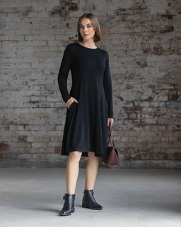 A woman with short brown hair and a neutral expression stands before a textured brick wall, wearing the Vassalli Lotus Merino Dress in black with black ankle boots and holding a burgundy handbag.