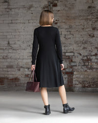 A woman wearing the Vassalli Lotus Merino Dress in black and black ankle boots faces a white brick wall, holding a maroon handbag in her left hand. Her straight hair is cut to shoulder length.