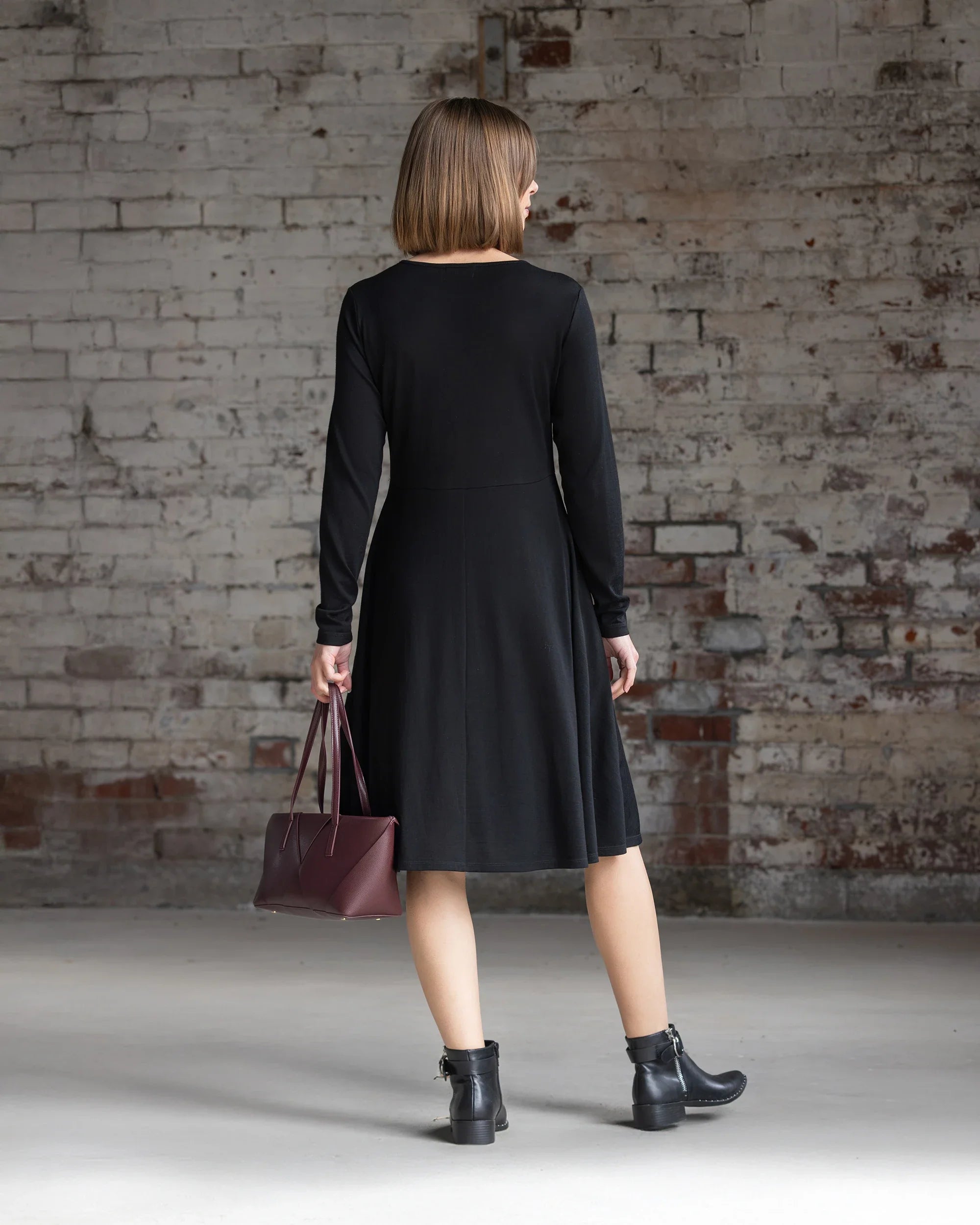 A woman wearing the Vassalli Lotus Merino Dress in black and black ankle boots faces a white brick wall, holding a maroon handbag in her left hand. Her straight hair is cut to shoulder length.