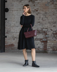 A woman wearing the Vassalli Lotus Merino Dress in black and black ankle boots stands indoors by a weathered brick wall, holding a maroon handbag and gazing thoughtfully to the side.