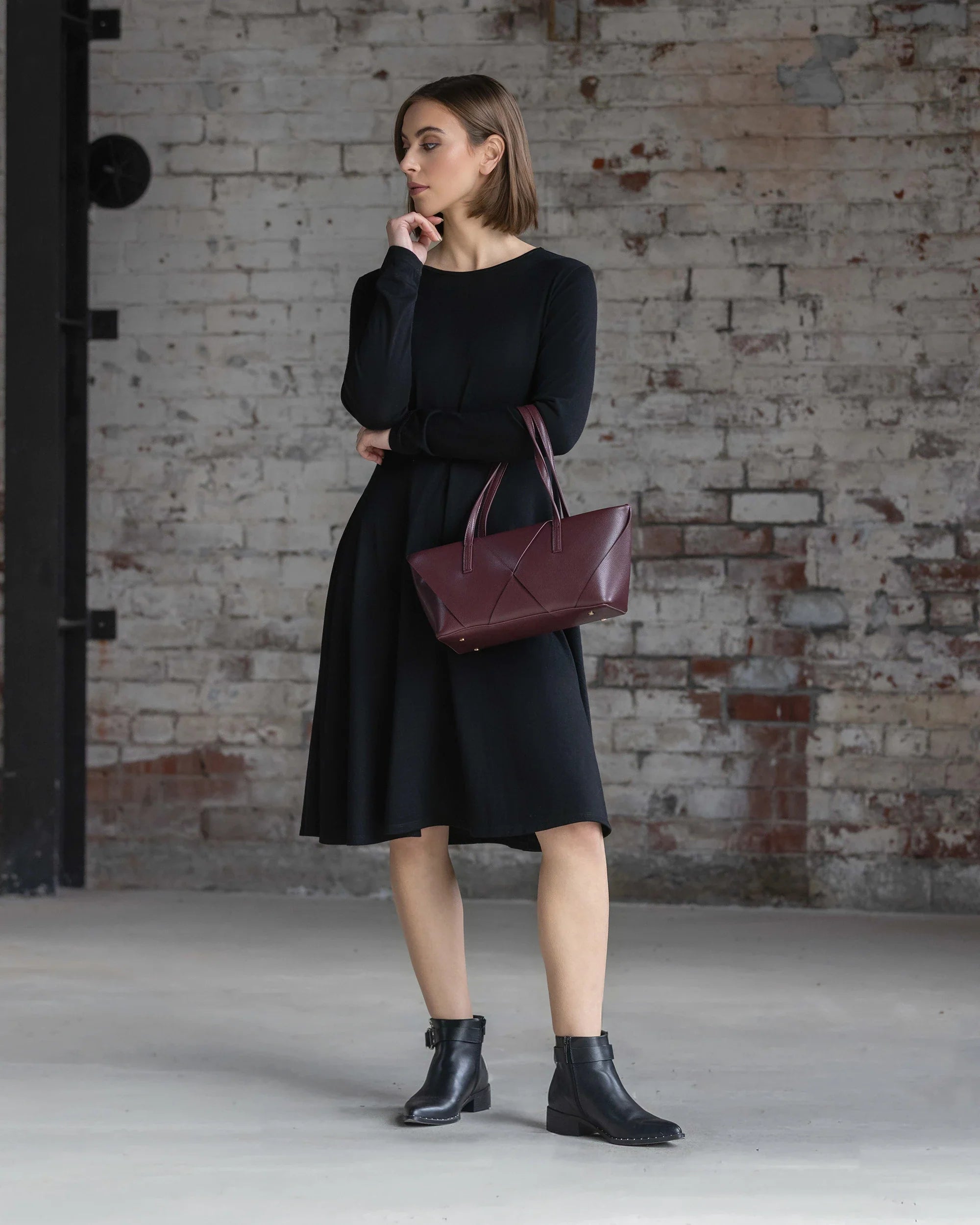 A woman wearing the Vassalli Lotus Merino Dress in black and black ankle boots stands indoors by a weathered brick wall, holding a maroon handbag and gazing thoughtfully to the side.