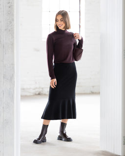A woman smiles and looks to the side in a bright, minimalist room, wearing the Vassalli Carrie Merino Skirt in black, paired with a maroon long-sleeve top, dark ankle boots, and holding a small matching handbag.