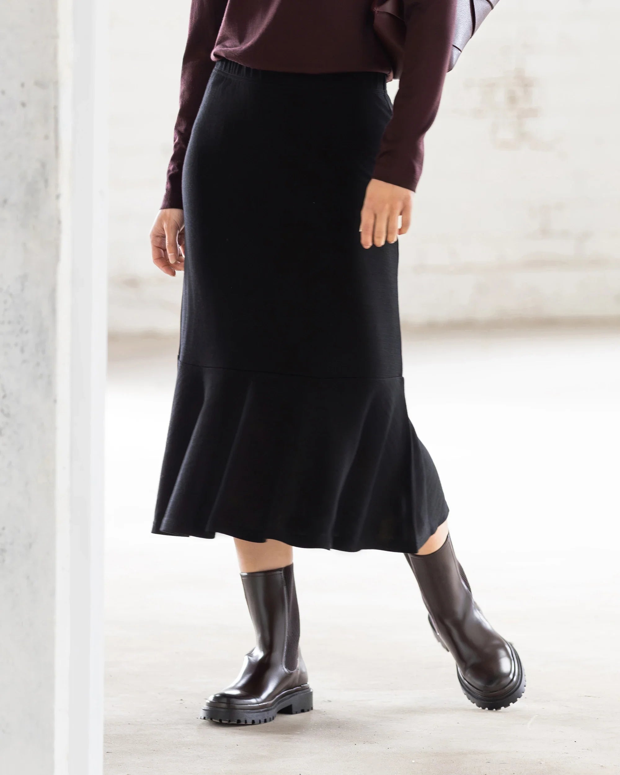 A person stands indoors with one knee slightly bent, wearing the Vassalli Carrie Merino Skirt in black, a dark long-sleeve top, and black ankle boots. The softly lit background is minimal.
