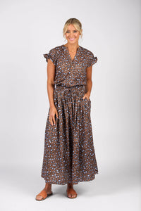 A woman with her blonde hair in a loose, low updo smiles against a white background. She wears the Vassalli Maxi Skirt - Nala in brown and blue leopard print with an elastic waistband, styled with short ruffled sleeves and tan sandals.