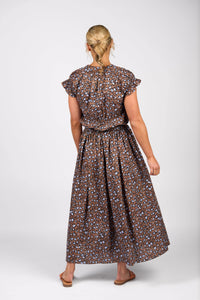 A woman faces away, wearing the Vassalli Maxi Skirt - Nala in a brown, blue, and black print. The skirt is cotton with an elastic waistband and a flowing silhouette. She pairs it with flat sandals, her blonde hair pulled back against a plain white background.