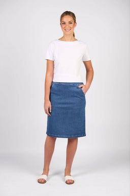 A woman stands smiling against a plain background, wearing the Vassalli Terra Skirt in Blue Hue Denim with a white short-sleeve top and open-toe sandals, one hand in her pocket, showcasing a relaxed casual style.