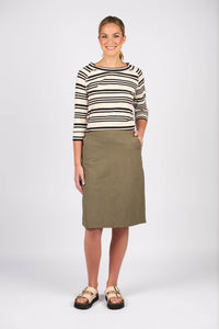 A woman smiles, wearing a black and white striped long-sleeve shirt, the Vassalli Belen Skirt - Army, and beige sandals. One hand is in her skirt pocket as she stands against a plain white background.