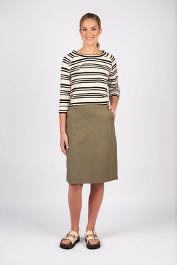 A woman smiles, wearing a black and white striped long-sleeve shirt, the Vassalli Belen Skirt - Army, and beige sandals. One hand is in her skirt pocket as she stands against a plain white background.