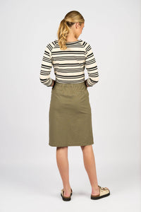 A woman with blonde hair in a ponytail stands facing away, wearing a black and white striped long-sleeve shirt, the Vassalli Belen Skirt - Army, and sandals against a plain light background.