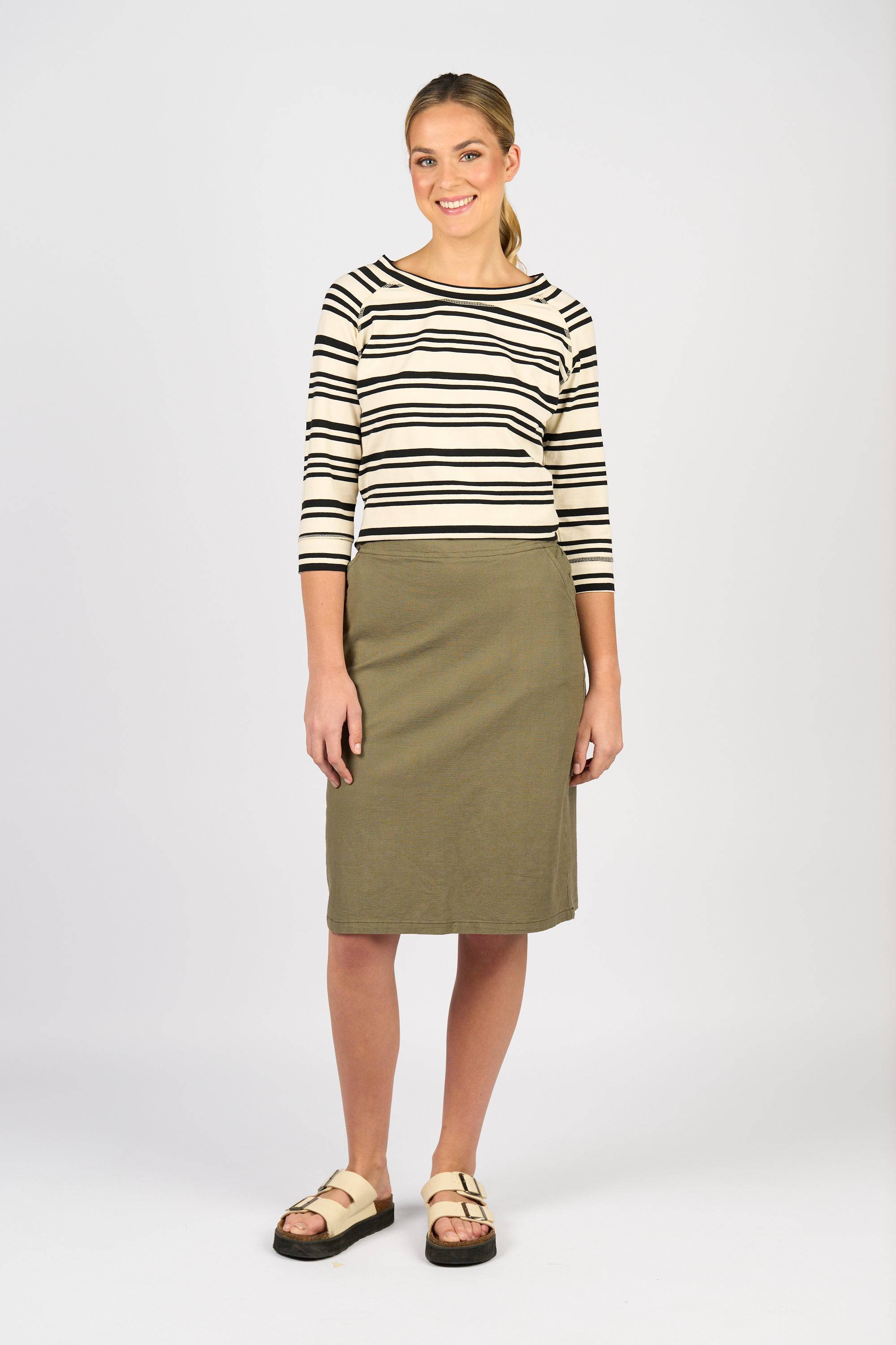 A woman smiles against a light grey background, wearing a cream and black striped 3/4 sleeve top, the Vassalli Belen Skirt - Army, and beige buckle sandals.