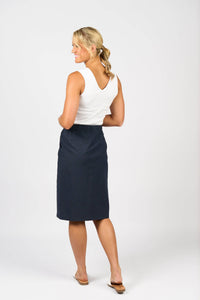 A woman with blonde hair in a ponytail stands smiling and facing away, wearing a white sleeveless top, Vassalli's Belen Skirt - Ink, and sandals against a plain white background.