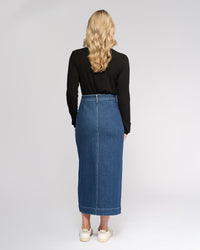 A woman with long, wavy blonde hair stands facing away, wearing a black long-sleeve top, white sneakers, and the Vassalli Rook Skirt - Dark Blue against a plain light background.