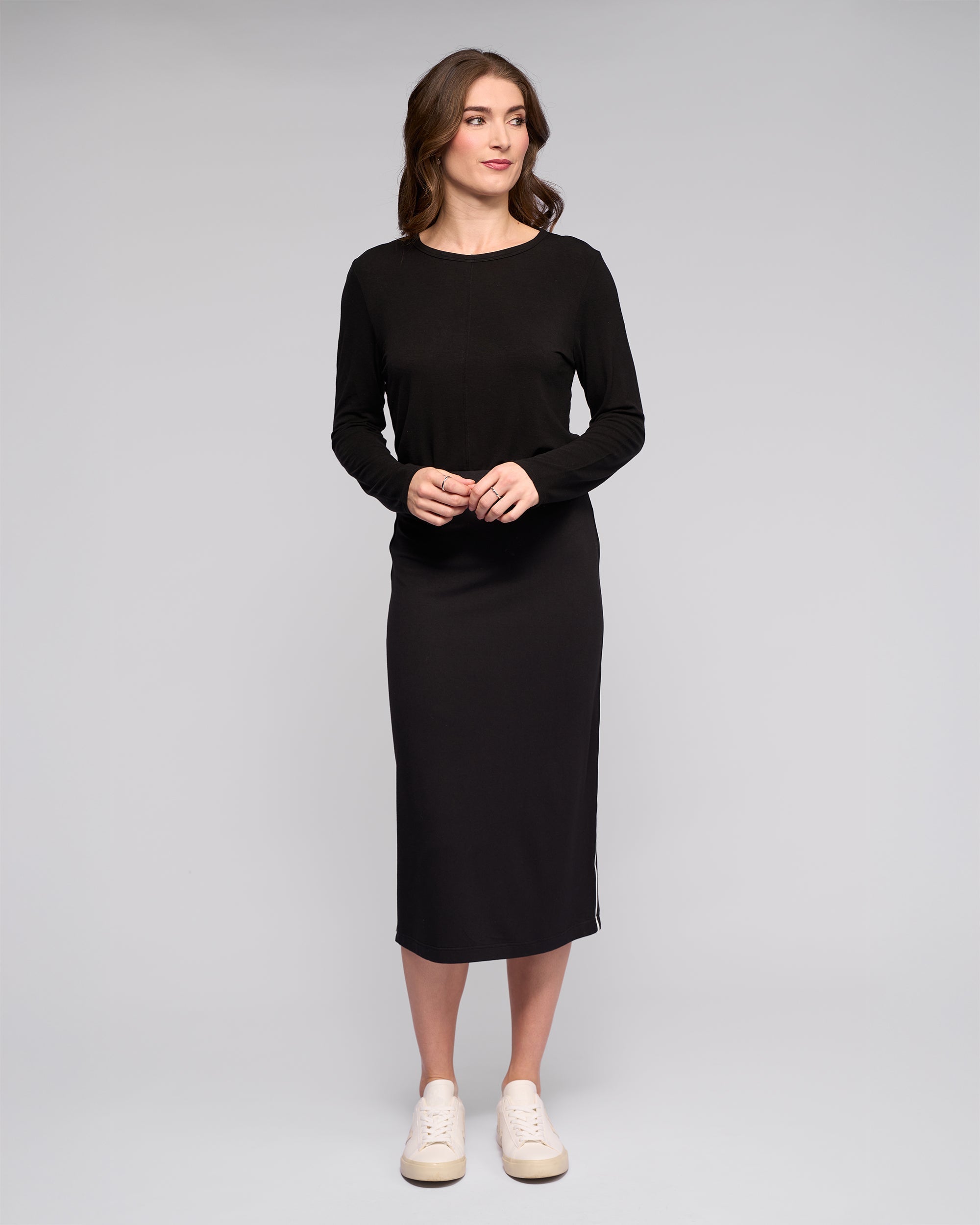 A woman with light brown hair stands against a plain light gray background, wearing the Vassalli Maple Skirt in black. She pairs it with white sneakers and a long-sleeved top, looking slightly to the side with her hands loosely clasped.
