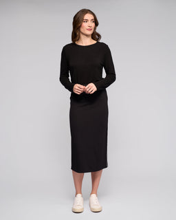 A woman with light brown hair stands against a plain light gray background, wearing the Vassalli Maple Skirt in black. She pairs it with white sneakers and a long-sleeved top, looking slightly to the side with her hands loosely clasped.