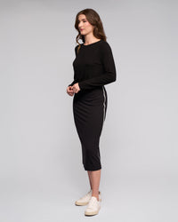 A woman with wavy brown hair stands sideways against a plain background, wearing Vassalli’s Maple Skirt – Black, featuring white side stripes and a stretchy midi fit. She pairs it with a long-sleeved black top and white sneakers, hands together in front.