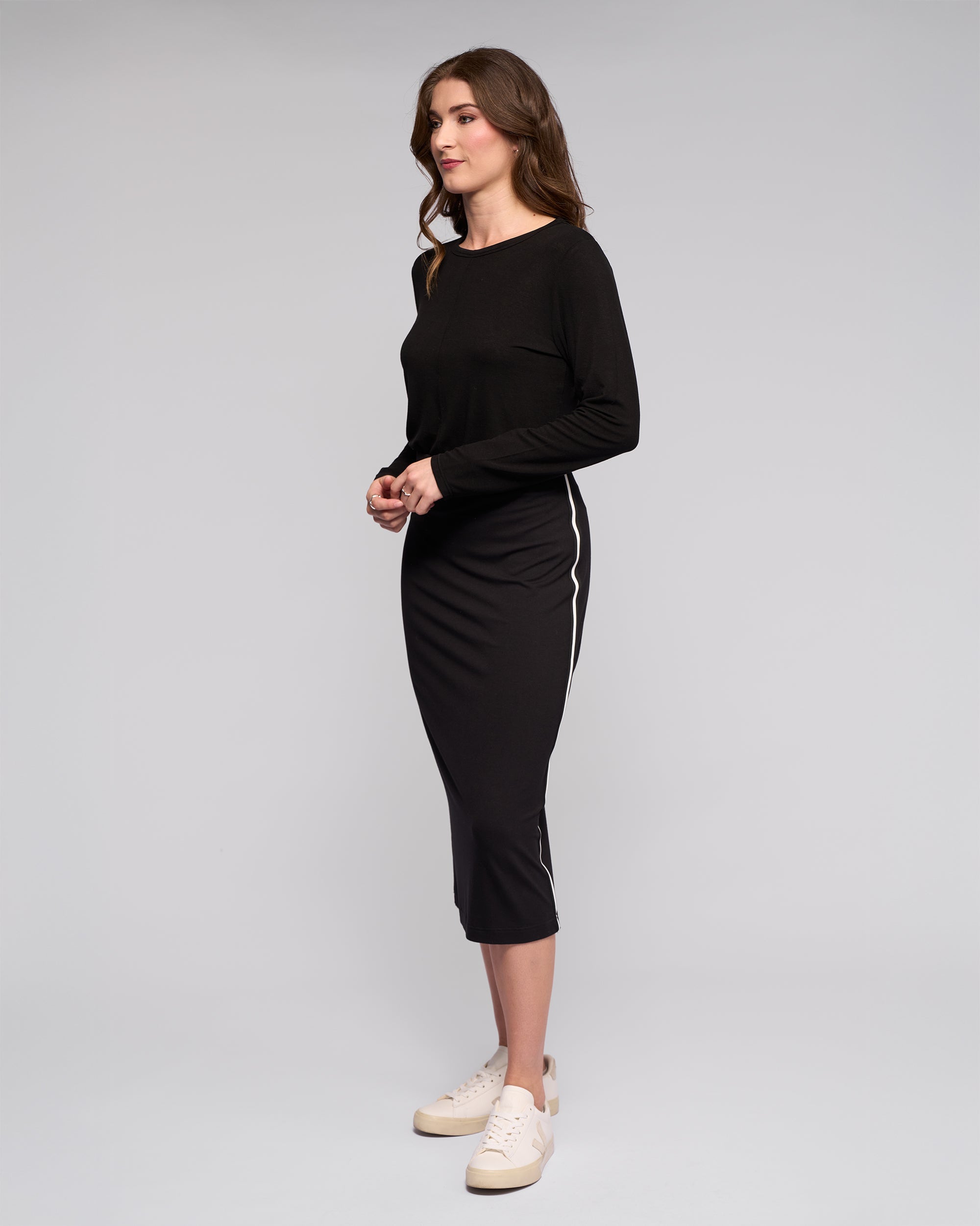 A woman with wavy brown hair stands sideways against a plain background, wearing Vassalli’s Maple Skirt – Black, featuring white side stripes and a stretchy midi fit. She pairs it with a long-sleeved black top and white sneakers, hands together in front.