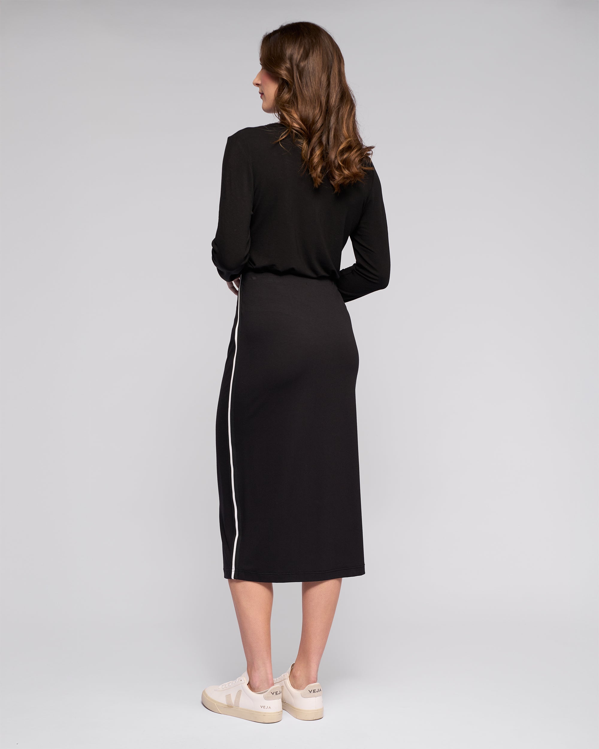 Woman stands with her back to the camera in a Vassalli Maple Skirt in black, paired with cream sneakers. The skirt features a white side stripe, stretchy fabric, and is photographed against a plain light grey background.