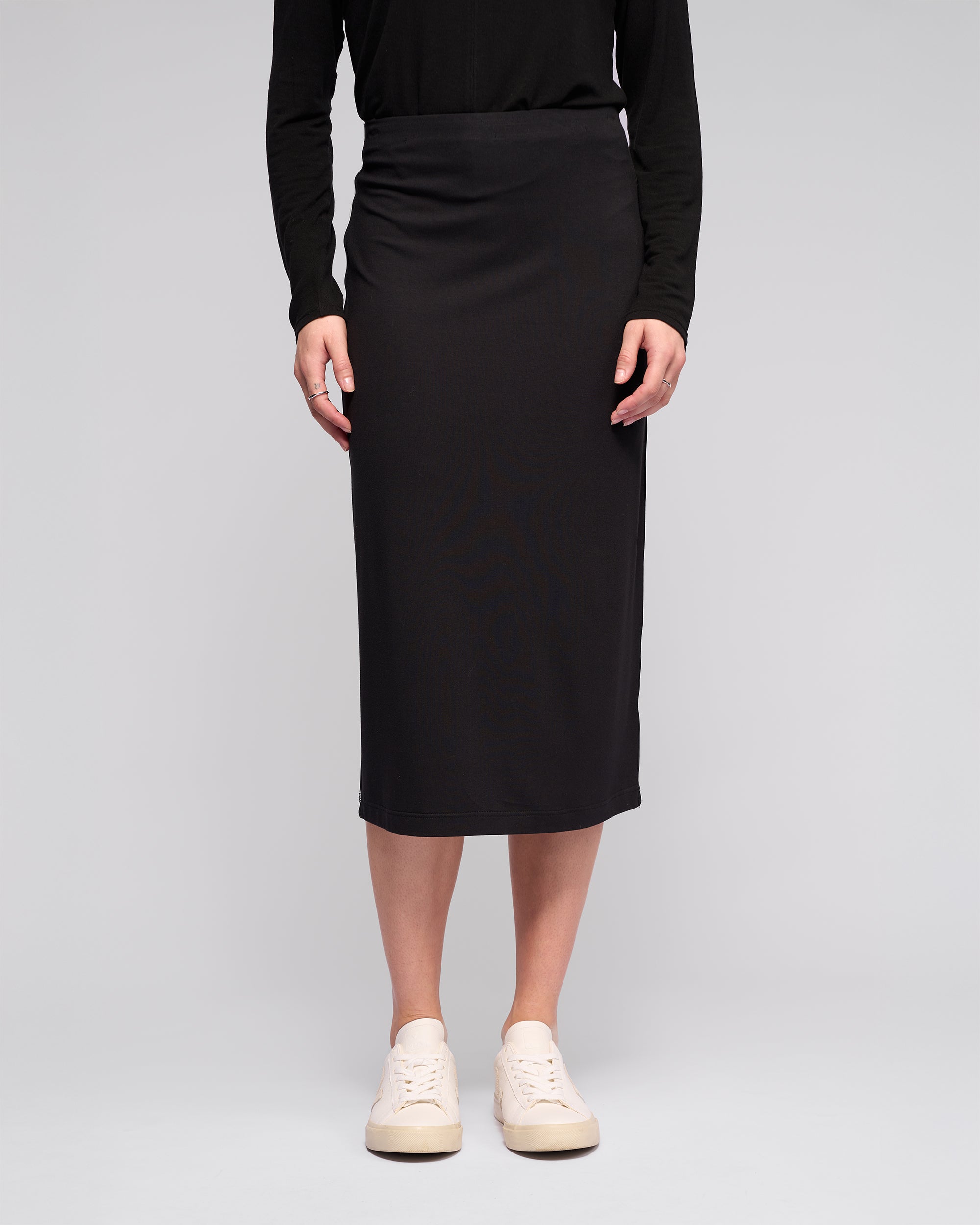 A person models the Vassalli Maple Skirt - Black, paired with a black long-sleeve top and white sneakers, against a plain light gray background. The upper body is partially out of frame. Perfect for stylish travel wear.