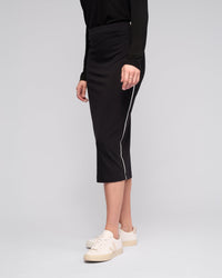 A person wearing the Vassalli Maple Skirt - Black, a black long-sleeve top, and white sneakers stands against a plain light background. The skirt is midi-length with stretchy fabric and a white side stripe. The person's face is not visible.