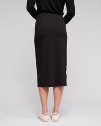 A person is seen from behind in the Vassalli Maple Skirt - Black, a stretchy midi skirt, paired with white Veja sneakers against a plain light background—an ideal stylish choice for travel.