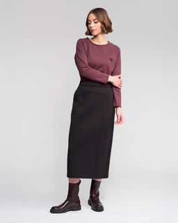 A woman poses against a plain background in the Vassalli Falcon Skirt - Black, paired with a long-sleeved maroon top featuring a heart graphic and dark brown ankle boots. She looks slightly to the side, one hand on her hip.