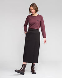 A woman in a long-sleeved maroon top with a small heart design, the Vassalli Falcon Skirt in black, and dark brown ankle boots stands against a plain light gray background.