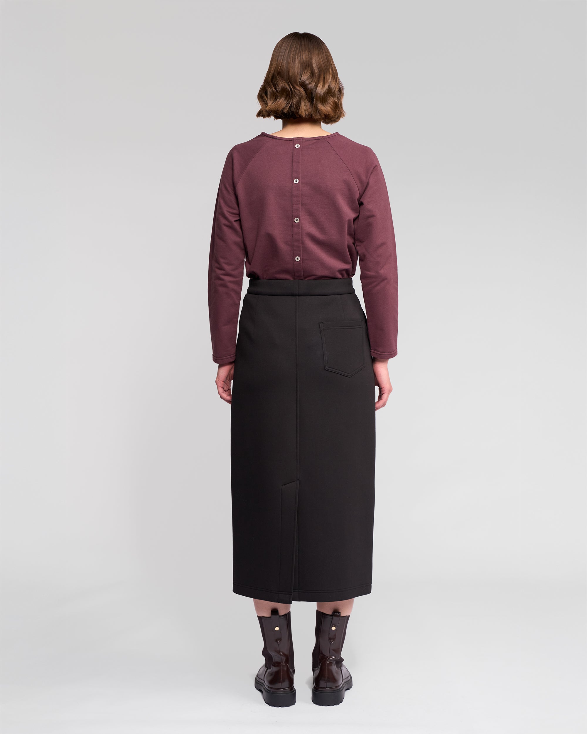 A person with short brown hair stands facing away in a mauve long-sleeve top and the Vassalli Falcon Skirt in Black, a high-waisted, winter-weight midi skirt with a back pocket, paired with black ankle boots against a neutral background.