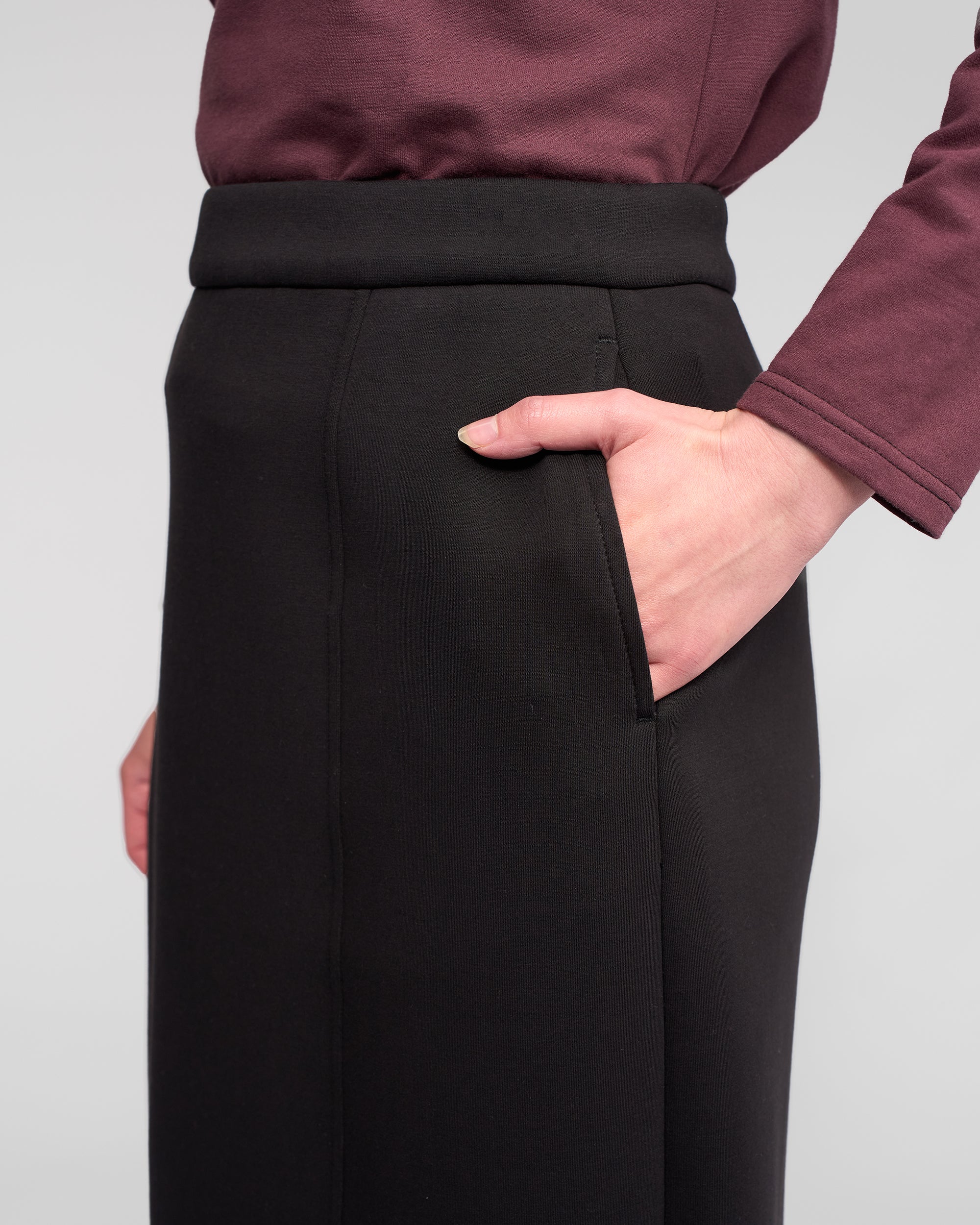A model wears the Vassalli Falcon Skirt in black, a high-waisted stretch knit midi with pockets, shown from the waist down against a plain background.