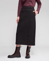 A person wears the Vassalli Falcon Skirt in black, paired with a maroon long-sleeve top and brown ankle boots, standing with one hand in their pocket against a plain light background.