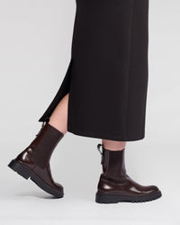 Someone stands against a plain light background wearing the Vassalli Falcon Skirt in black, a winter-weight stretch knit skirt with a side slit, paired with dark brown chunky-soled ankle boots.