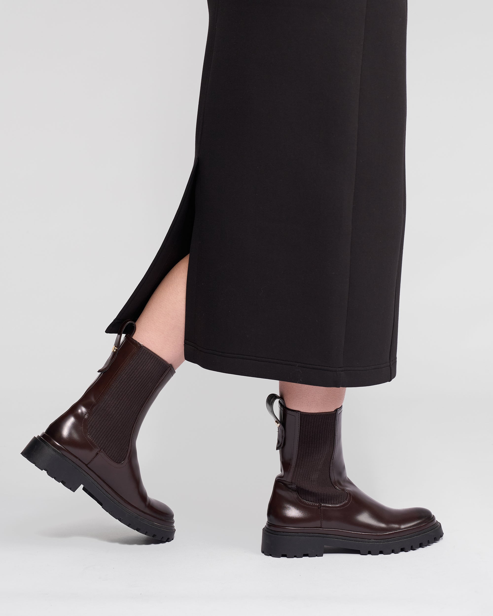 Someone stands against a plain light background wearing the Vassalli Falcon Skirt in black, a winter-weight stretch knit skirt with a side slit, paired with dark brown chunky-soled ankle boots.