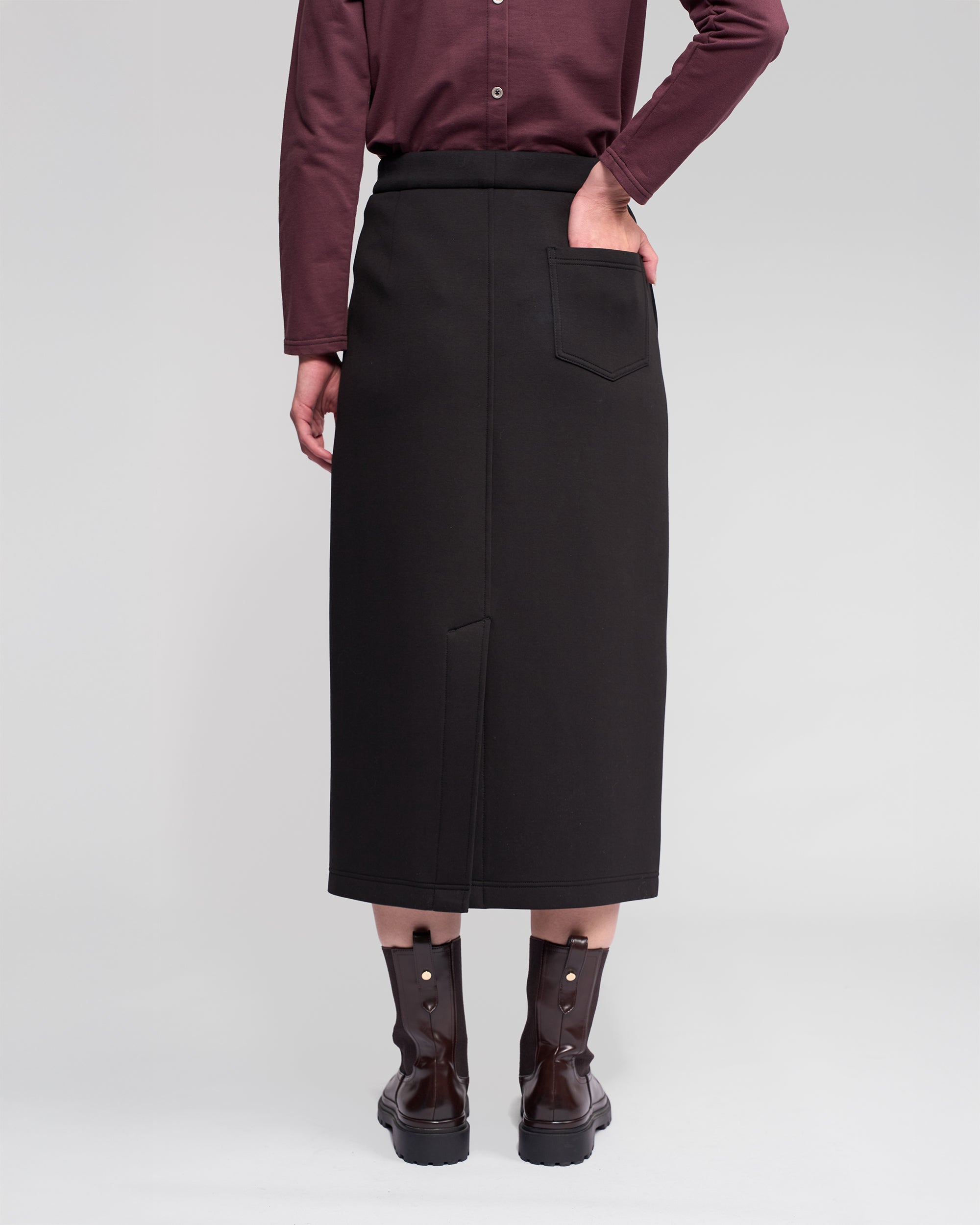 A person stands facing away, wearing a burgundy long-sleeve shirt, dark ankle boots, and the Vassalli Falcon Skirt in black with a back pocket. One hand rests in the skirt's pocket. The background is plain and light.
