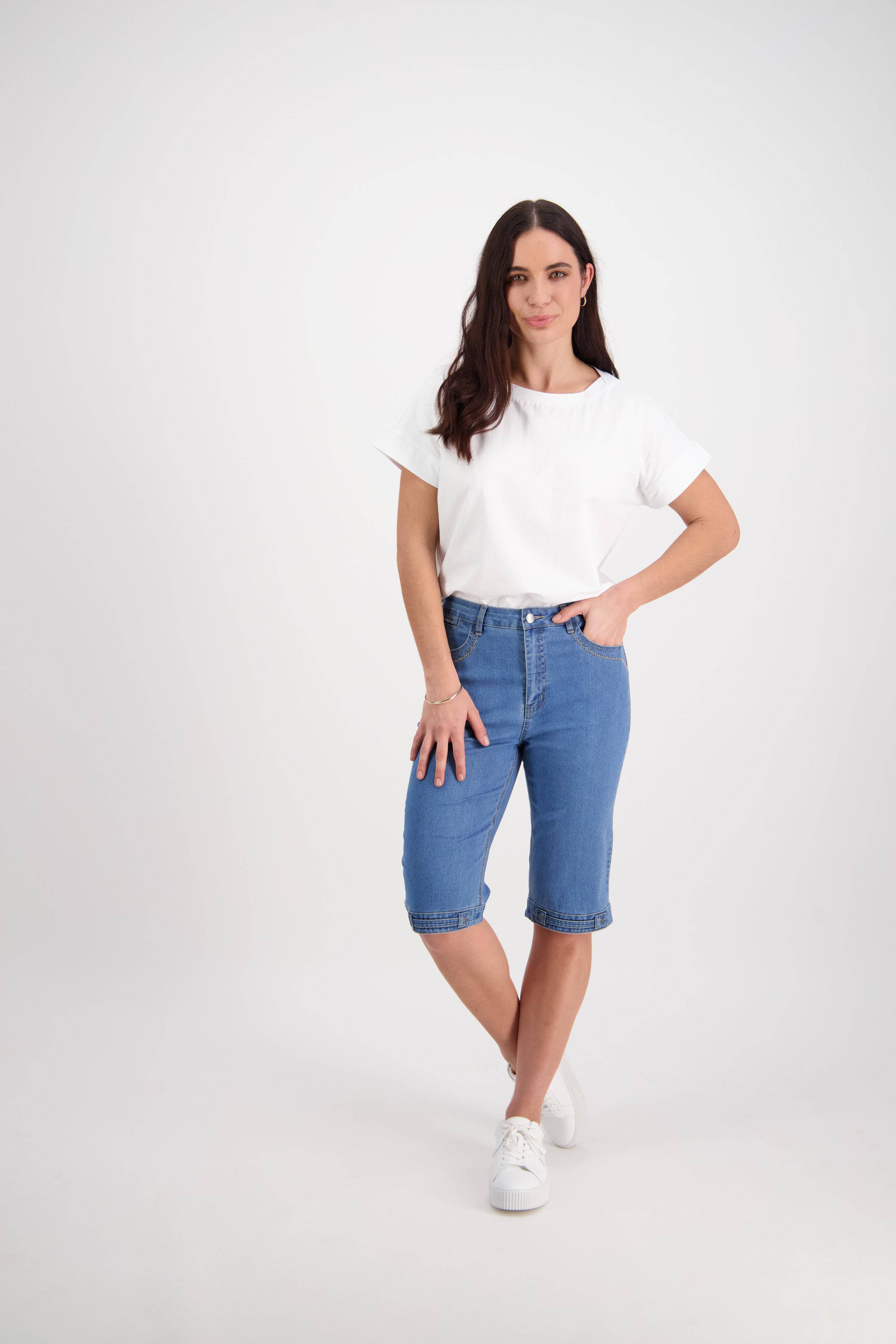 A woman stands against a white background wearing Vassalli's Custom Short - Light Denim, a stretch-fabric straight-leg blue denim short, paired with a white t-shirt and sneakers. She has long brown hair and smiles softly with one hand on her hip.