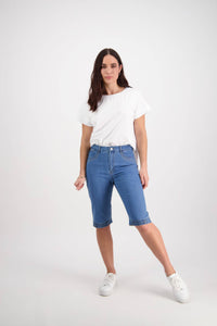 A woman with long brown hair and a neutral expression stands against a white background, wearing Vassalli's Custom Short - Light Denim, a white T-shirt, and white sneakers, with one leg slightly bent.