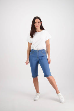 A woman with long brown hair and a neutral expression stands against a white background, wearing Vassalli's Custom Short - Light Denim, a white T-shirt, and white sneakers, with one leg slightly bent.