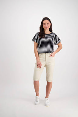 A woman stands against a white background, wearing a black and white striped short-sleeve top, Vassalli Custom Poplin Short in Sand with a cuffed hem, and white sneakers. She smiles gently with one hand in her pocket.