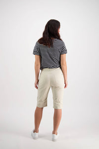 A woman with long brown hair faces away, wearing a black and white striped t-shirt, Vassalli Custom Poplin Short in Sand, and white sneakers against a plain white background.