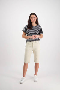 A woman with long brown hair smiles gently at the camera, wearing a black and white striped t-shirt, Vassalli Custom Poplin Short in Sand, and white sneakers against a plain white background.